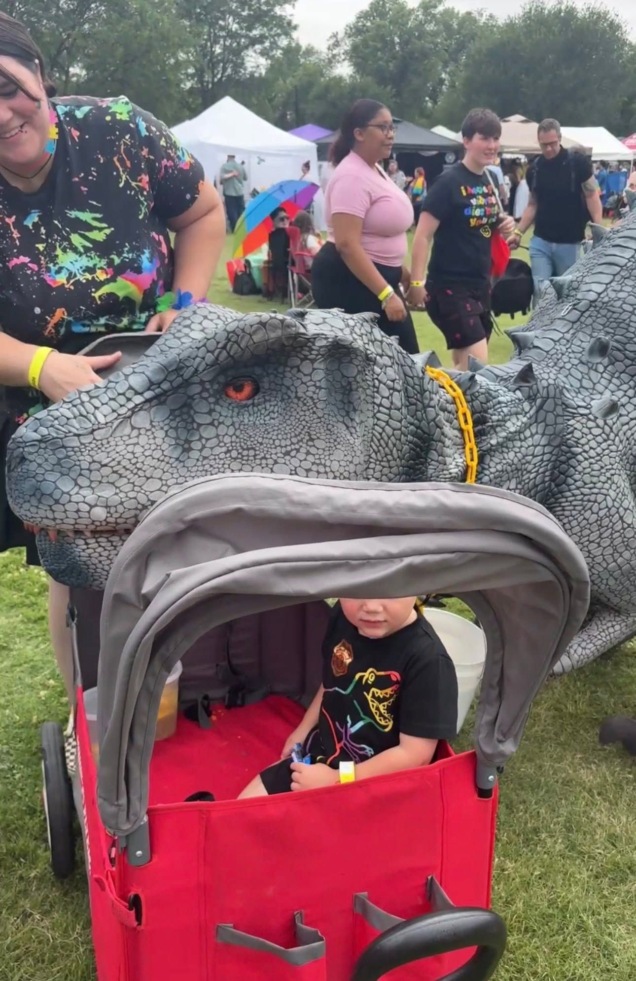 Person in a red stroller costume with a gray dinosaur head at an outdoor festival