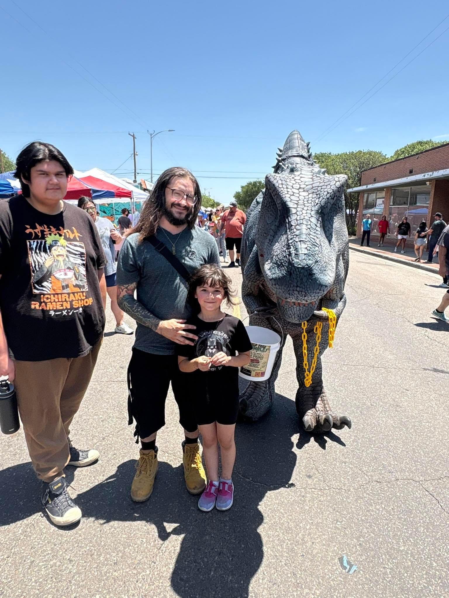 Three people posing beside a gray dinosaur mascot at an outdoor street festival.