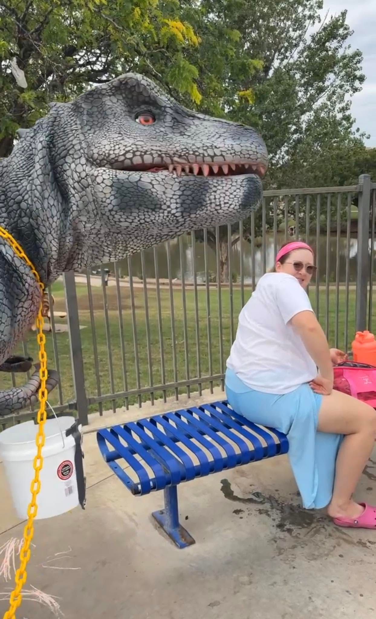 Person sitting on a blue bench beside a large dinosaur statue outdoors