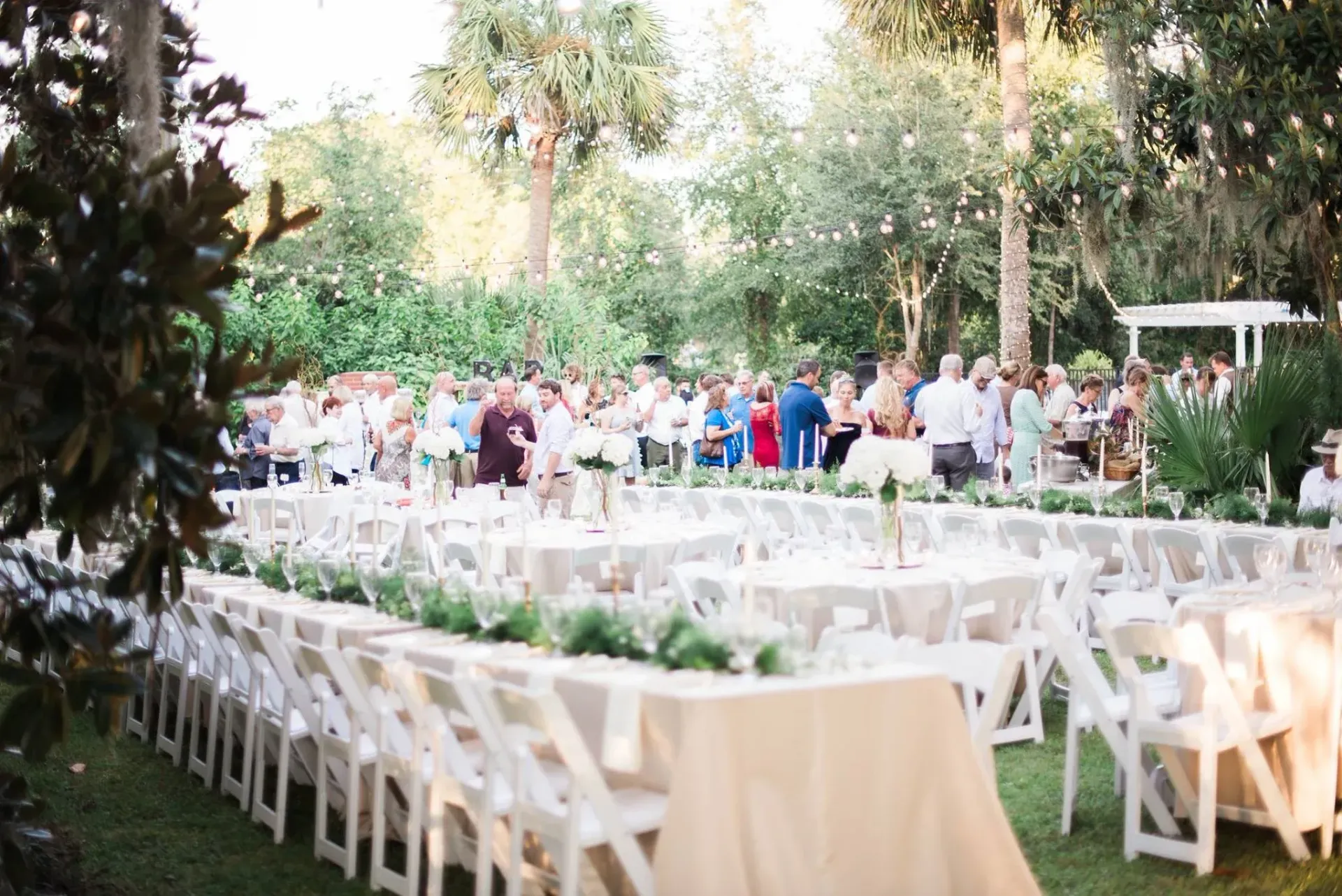 Outdoor wedding reception with long white tables and chairs set on a lawn, guests mingling in the background