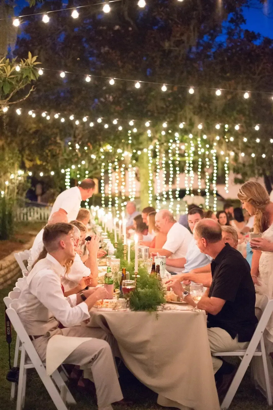 Outdoor dinner party under string lights, with guests seated at a long candlelit table at dusk
