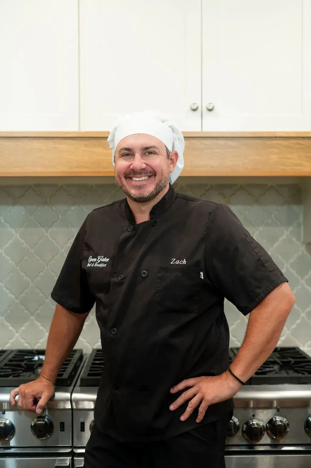 Chef in black uniform and hat standing in a kitchen, smiling with hands on hips.