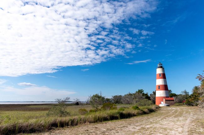 Red and white striped lighthouse beside a sandy path under a bright blue sky with clouds