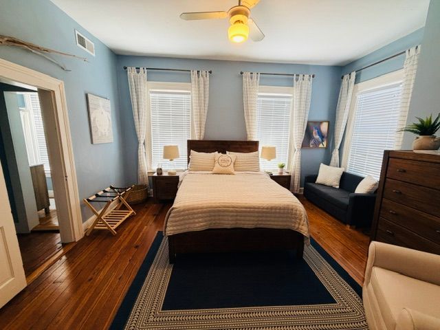 Bedroom with floral bedspread, wicker bench, blue rug, and twin windows with white blinds
