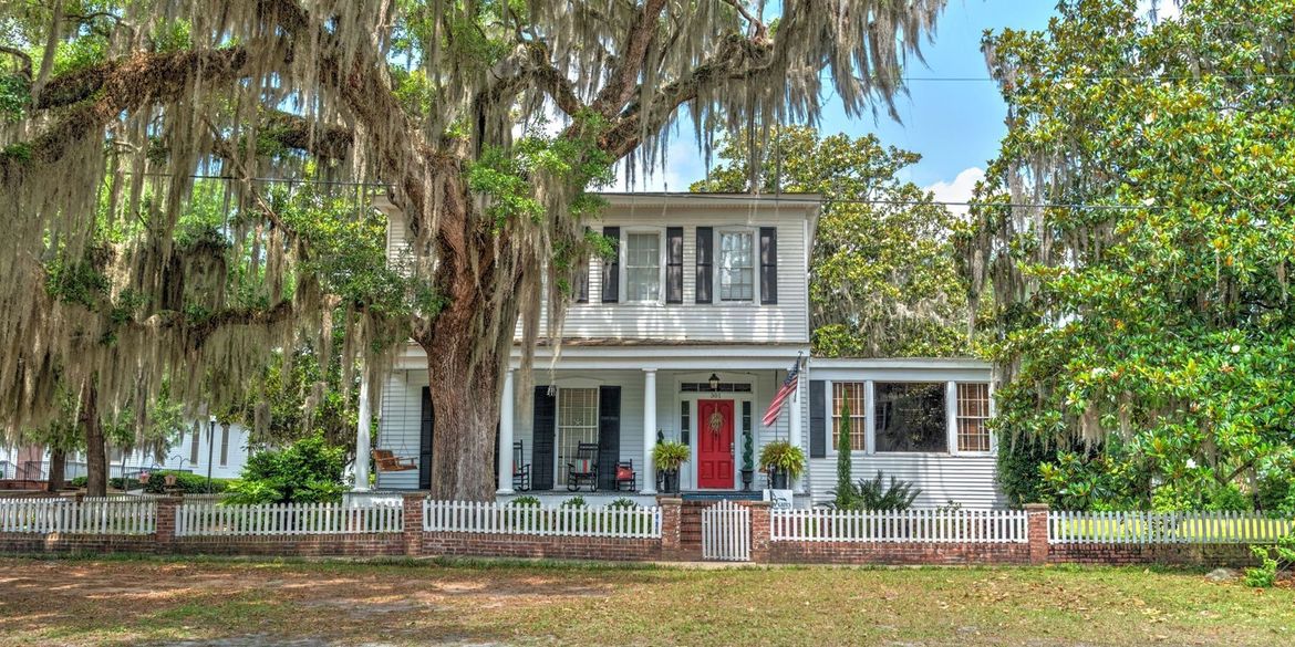 White historic house with porch, red front door, and a white picket fence under large trees