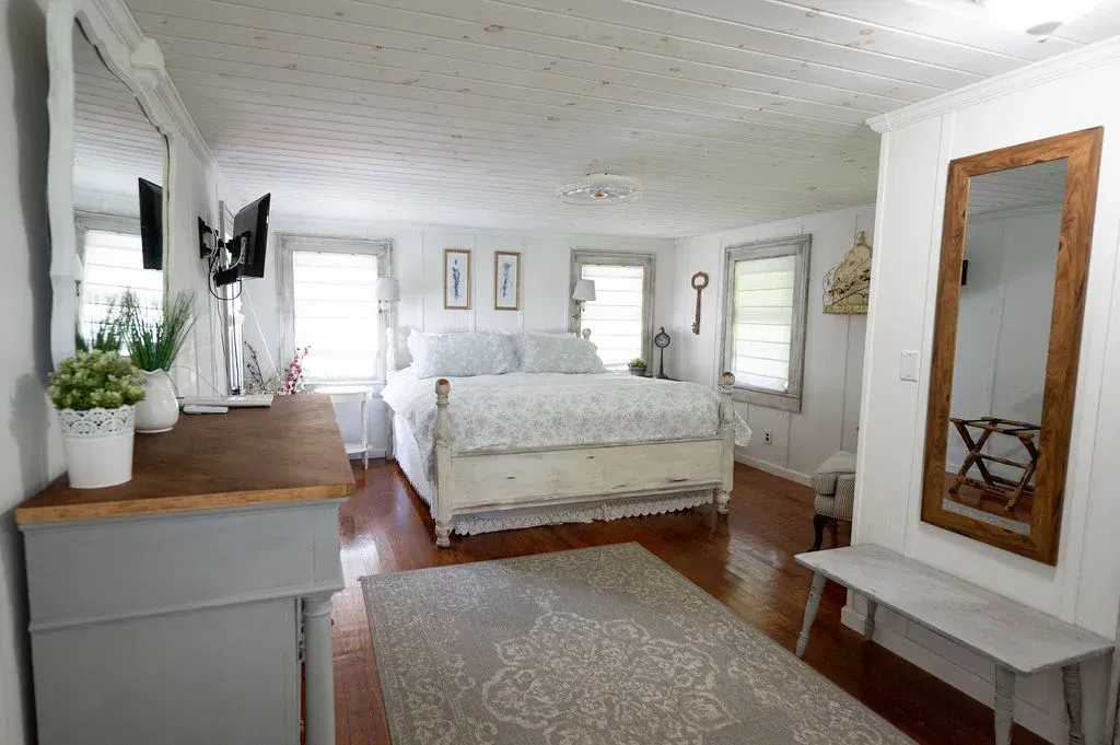 Bright white bedroom with a bed, dresser, mirror, windows, and wooden floorboards