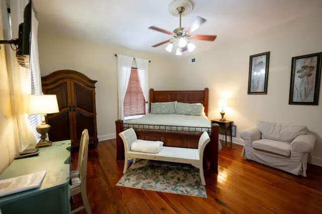 Spacious bedroom with a wooden bed, white bench, armoire, and ceiling fan on hardwood floors.