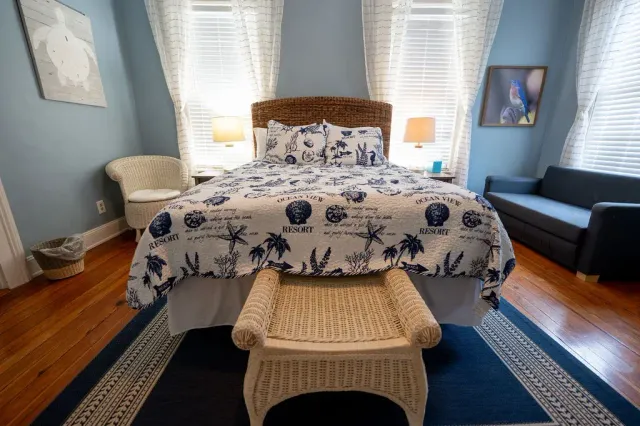 Bedroom with floral bedspread, wicker bench, blue rug, and twin windows with white blinds