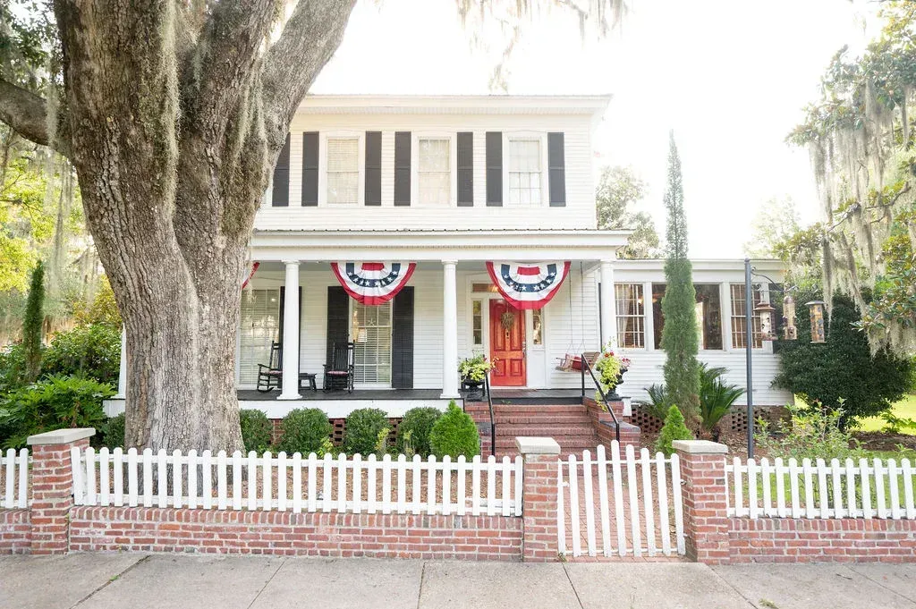 Historic white house with porch, red-white-blue bunting, and a white picket fence under large trees