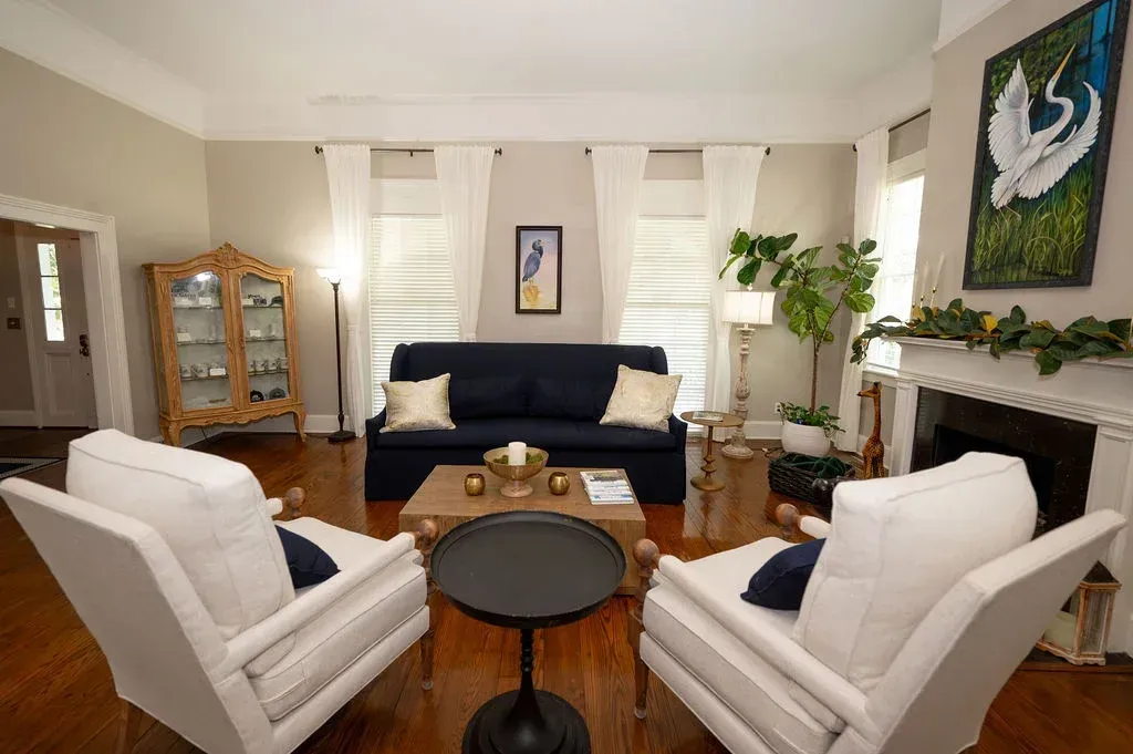 Bright living room with white chairs, dark sofa, wooden cabinet, and floral artwork.