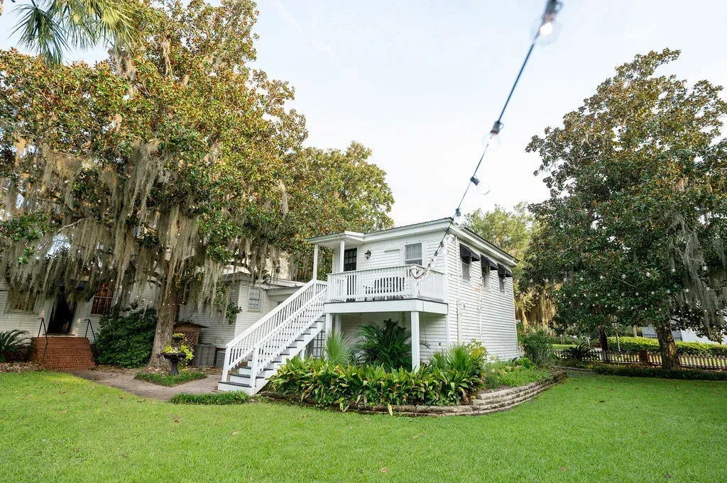 White elevated house with stairs, surrounded by lush green lawn and tall trees under a cloudy sky