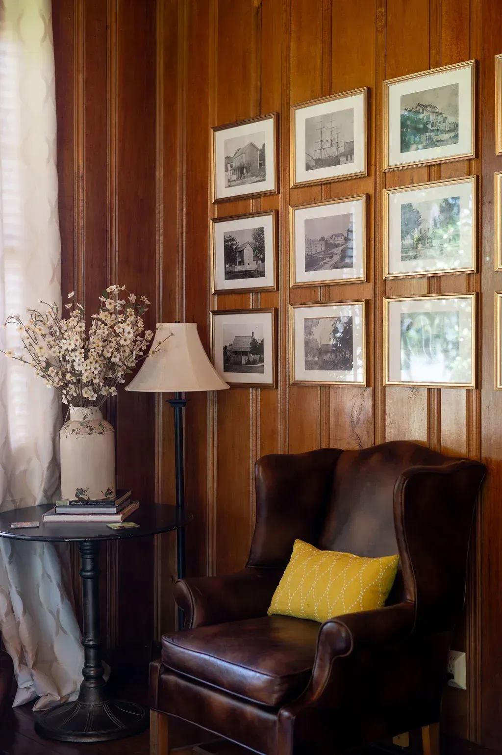 Cozy corner with a brown leather chair, yellow pillow, lamp, side table, and framed art on wood-paneled walls