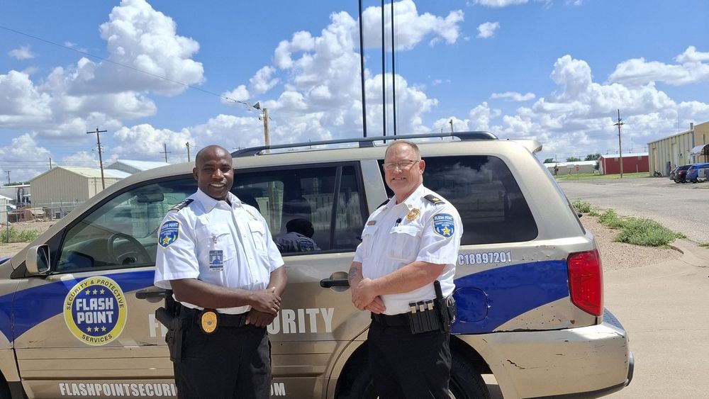 Two security officers stand beside a tan and blue patrol SUV under a cloudy sky.