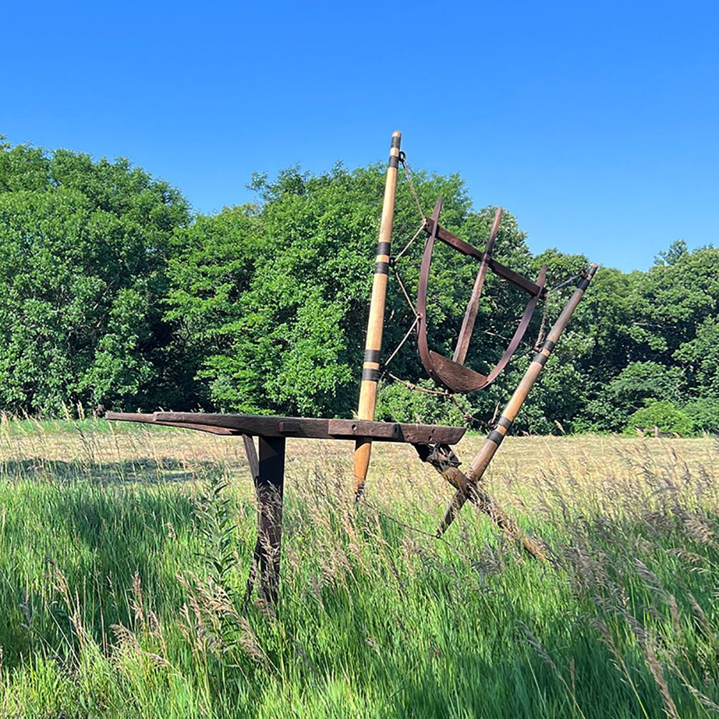 Weathered metal sculpture in grassy field, blue sky, trees in the background.