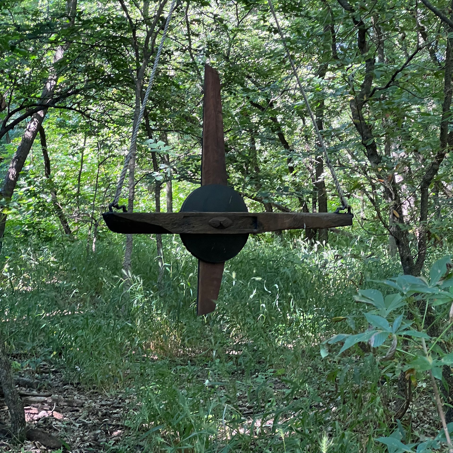 Rustic metal sculpture in a forest clearing. The sculpture resembles a cross with rusty blades.