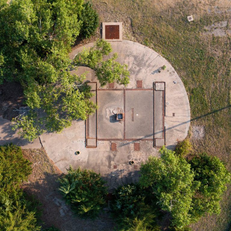 Overhead view of a concrete military installation with a square center and circular exterior, surrounded by trees and grass.