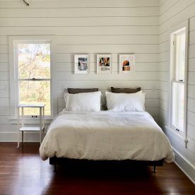 Bedroom with a bed, artwork, two windows, and dark wood floors. White walls and bedding.