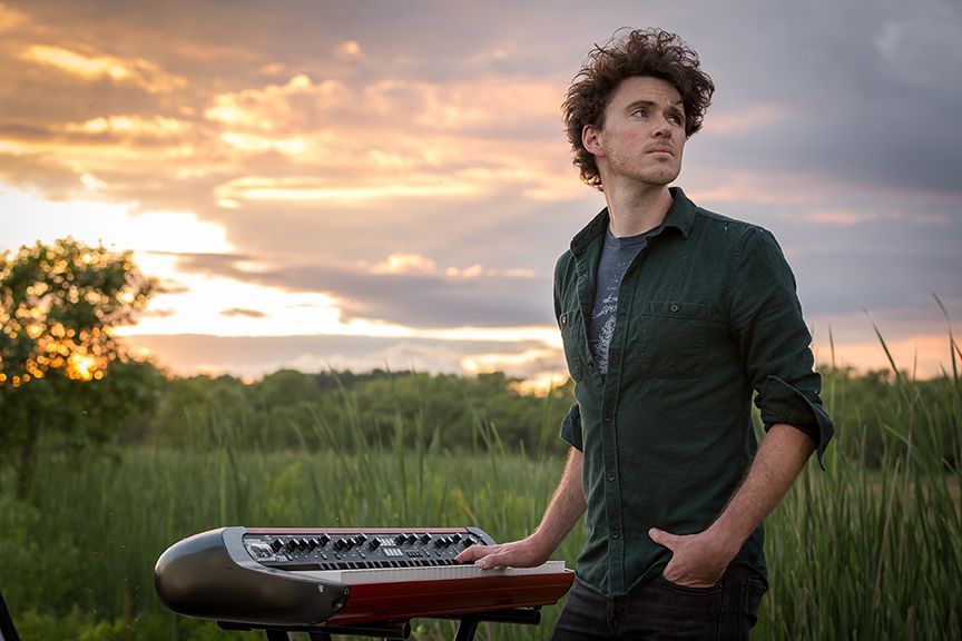 A young man is standing next to a keyboard in a field at sunset.