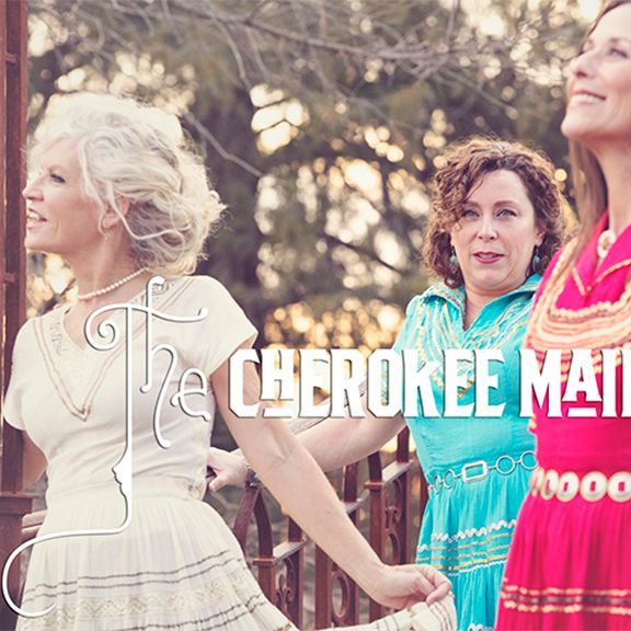 The Cherokee Maid: Three women in colorful dresses pose outdoors, smiling, and looking up.