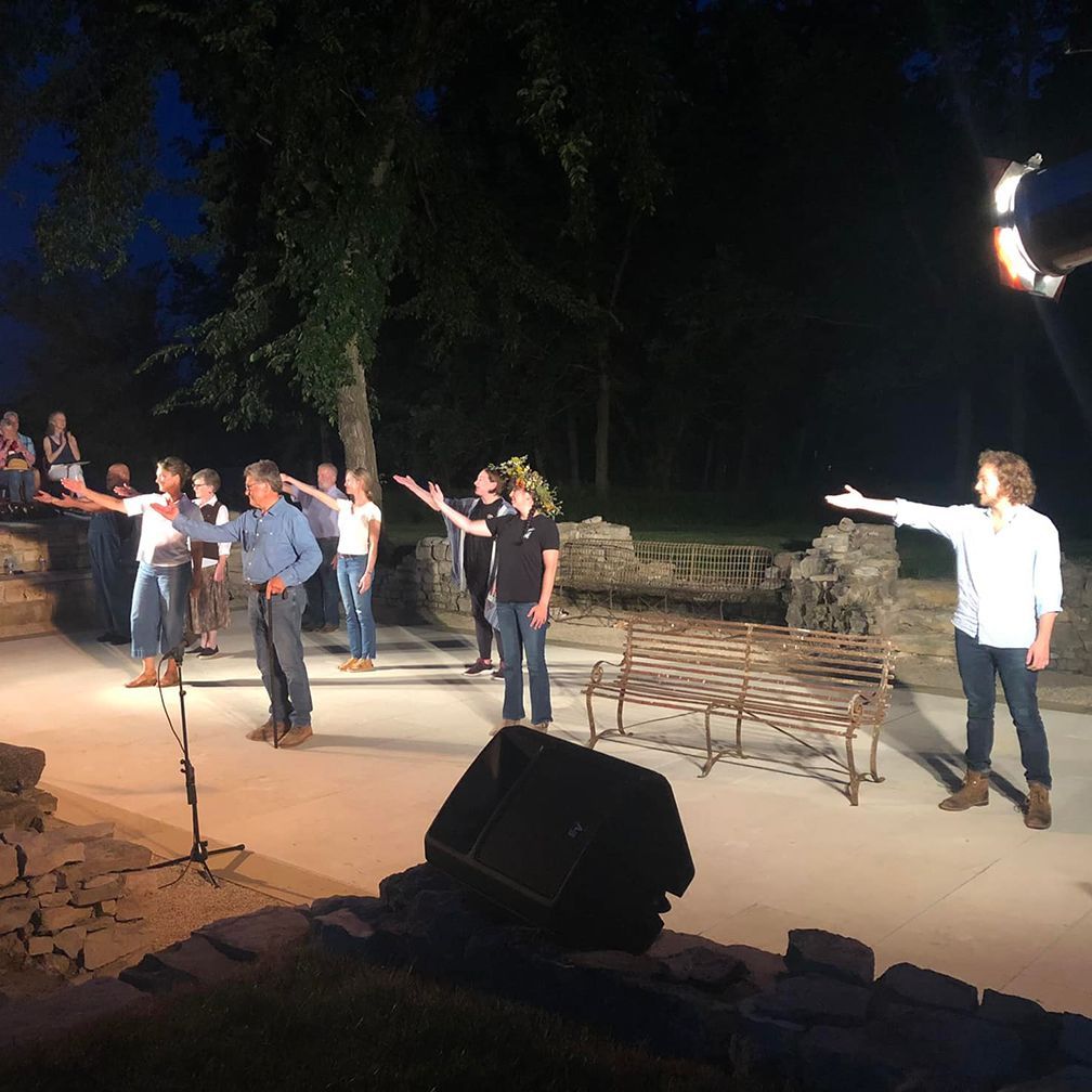 People on stage with arms outstretched, performing at night. Stone backdrop, benches, and trees.