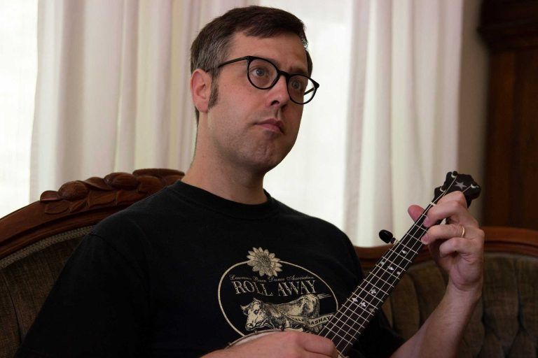 Man with glasses plays a banjo, sitting on an ornate sofa. He wears a black t-shirt indoors.