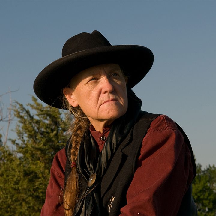 Woman in cowboy hat, looking upwards. Outdoors, wearing a black vest and red shirt.