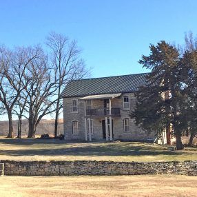 Two-story stone house with a balcony under a clear blue sky, surrounded by trees and a stone wall.