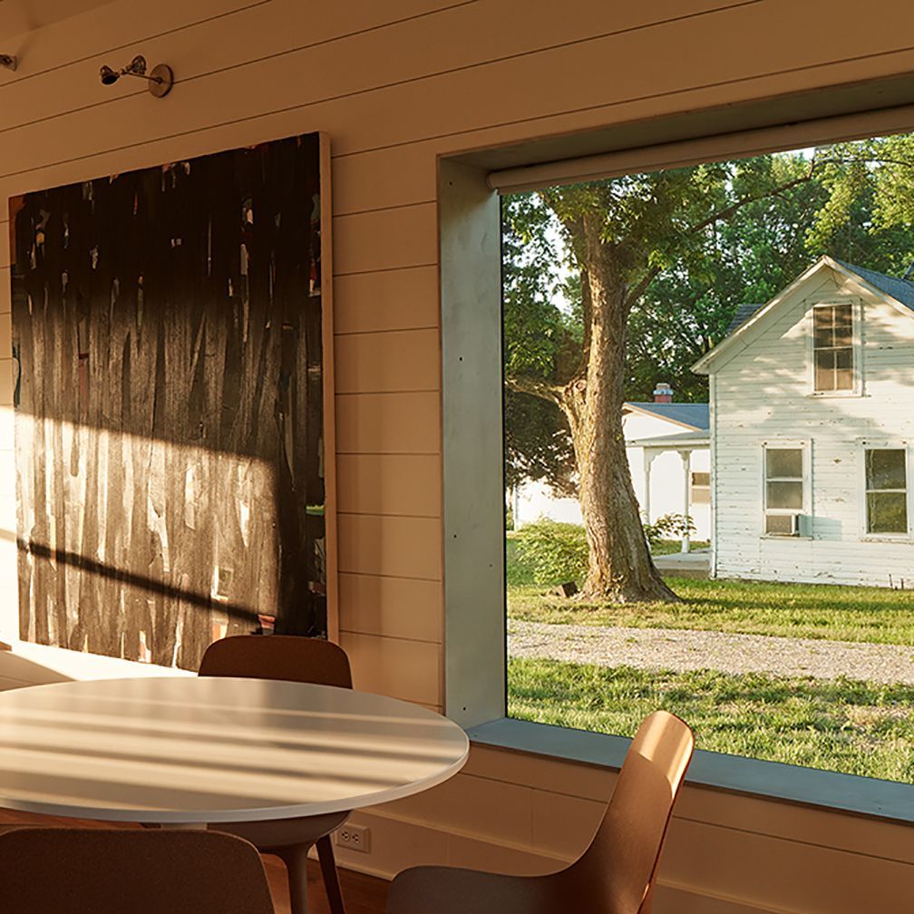 Interior view with round table, chairs, and window overlooking white house and tree.