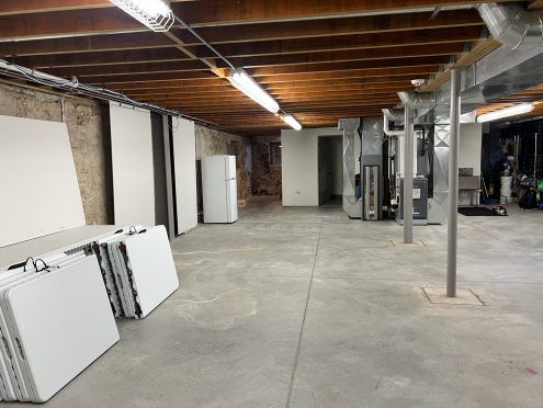 Basement with concrete floor, exposed ceiling beams, white folding tables, fridge, and HVAC units.