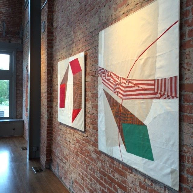 Two abstract art pieces hang on a red brick wall in a gallery. The pieces use red, green and white shapes.