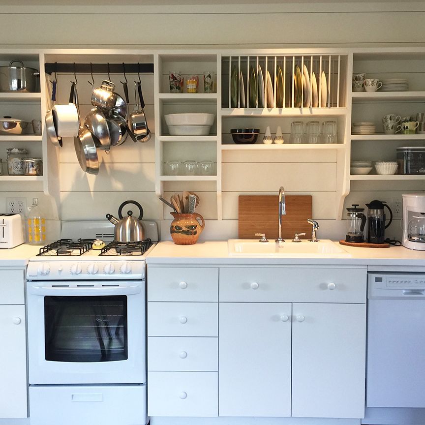 White kitchen with open shelving, stove, sink, and hanging pots.