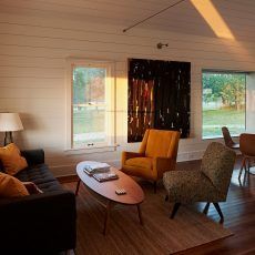 Living room with mid-century furniture, artwork, and large windows; bathed in warm sunlight.