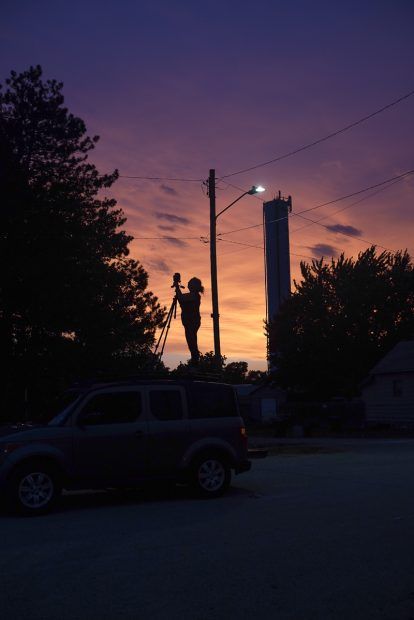 Silhouette of a person with a camera, a car, a tower, and trees against a colorful sunset.