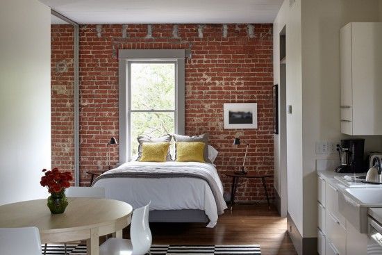 Bedroom with exposed brick wall, bed, small table, and open kitchen area.