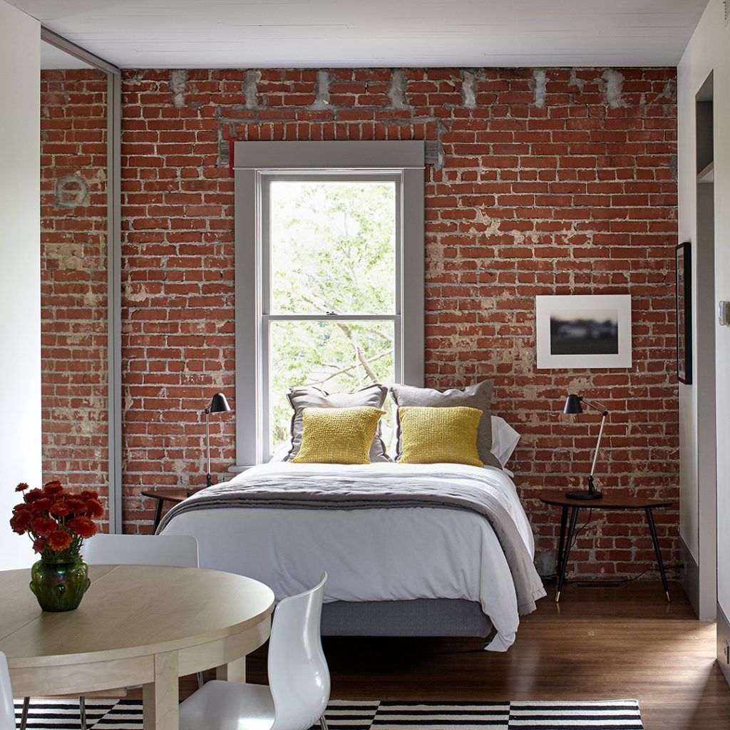 Bedroom with exposed brick wall, bed with gray bedding, yellow pillows, window, small side tables, and a dining table.
