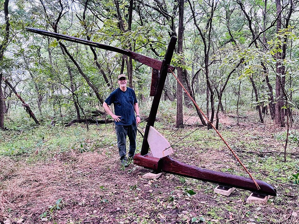 Man standing next to a large, rusty metal sculpture in a wooded area.