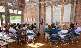 Woman giving a presentation in a brick-walled art gallery to a seated audience.
