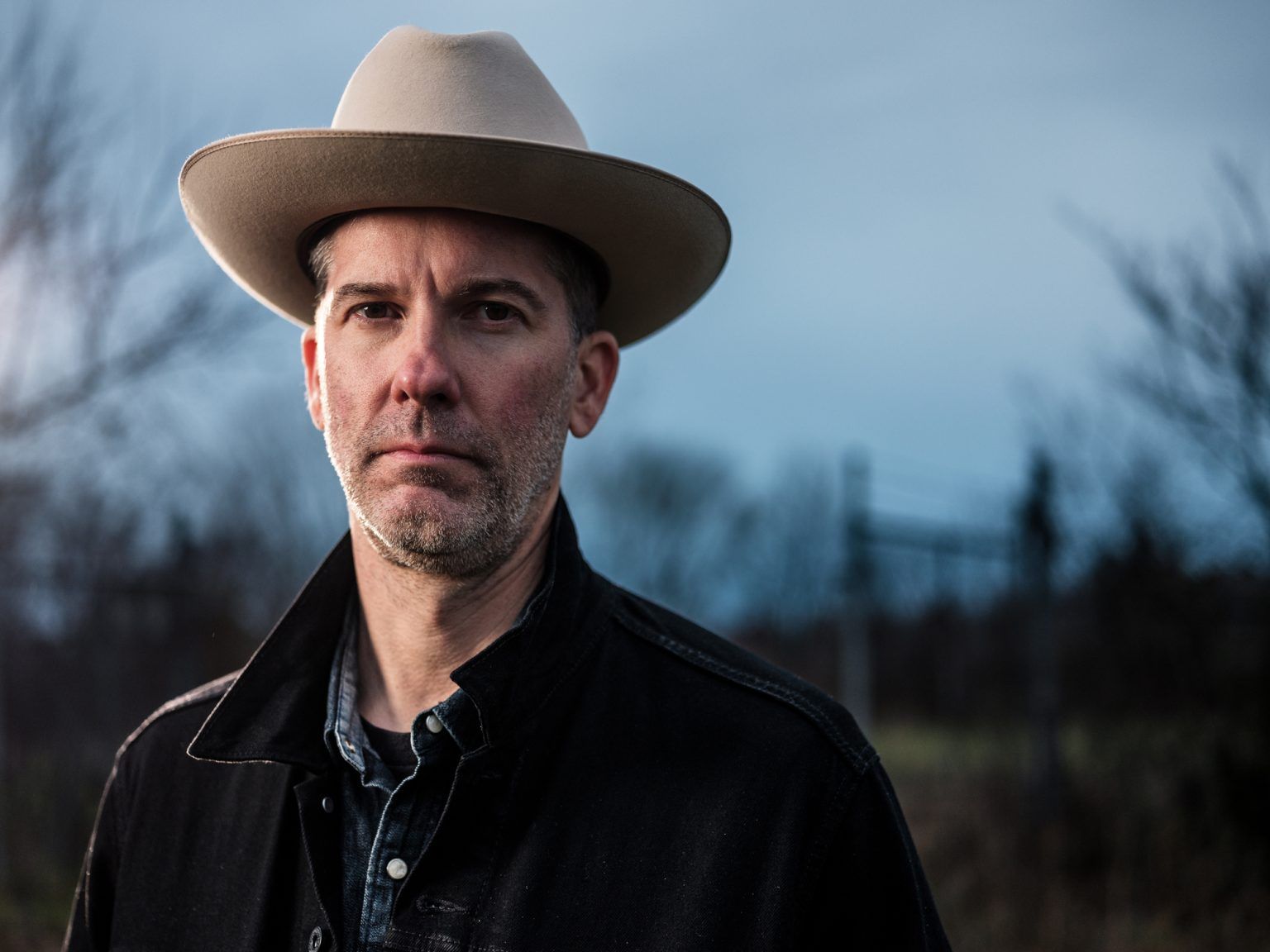 Man in a fedora and dark coat; outdoors with a serious expression; blurred background.