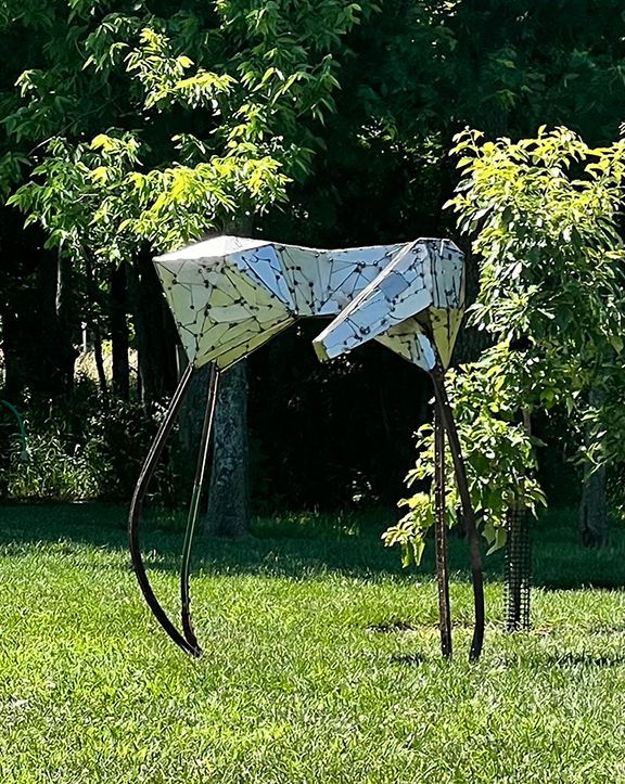 Metal sculpture of a geometric animal with long legs, in a grassy park, trees in the background.