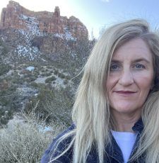 Blonde woman smiles in front of a red rock mountain with patches of snow. Blue sky and scrub brush.