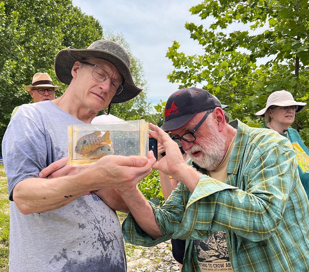 Two men examining an object in a small box, outdoors. One wears a hat