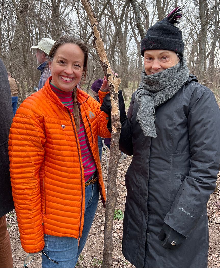 Two women smile, pose beside a small tree. One wears an orange jacket, the other a black coat and hat, outside.