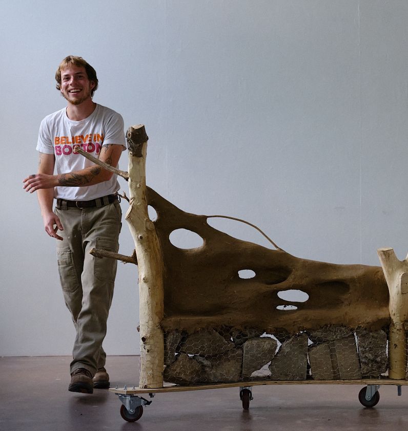 Man smiling next to a wood and stone art piece on wheels in a studio.