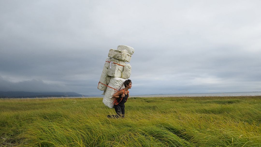 Man carries a towering stack of white bags across a green field under a cloudy sky.