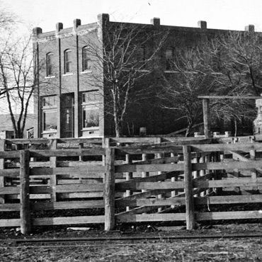 Two-story brick building with large windows, behind a wooden fence, bare trees, and a dirt ground.