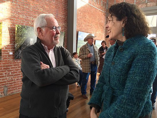 Man in gray sweater with arms crossed talking to a woman in a teal coat. Others in background.