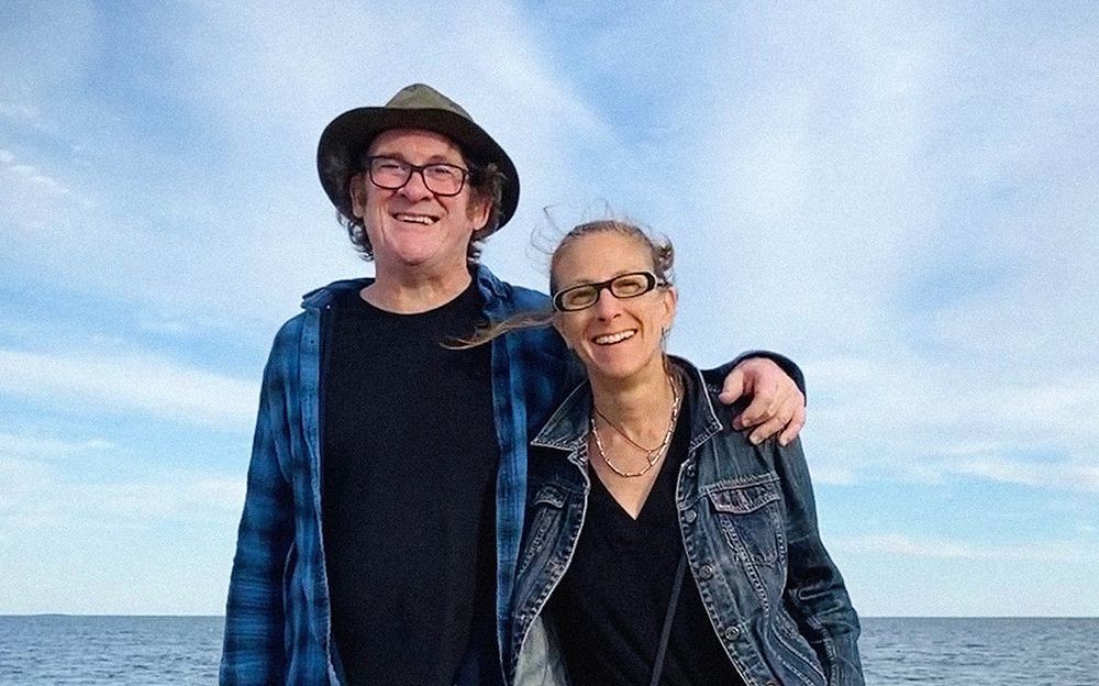 Man with hat and woman in glasses pose together smiling in front of a blue sky and water.