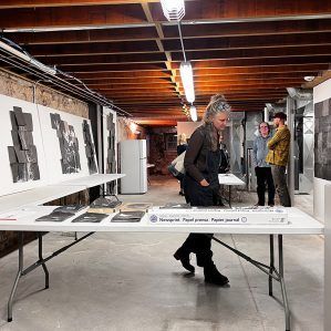 People at an art exhibit in a basement. Woman in dark clothing at a table, looking at art.