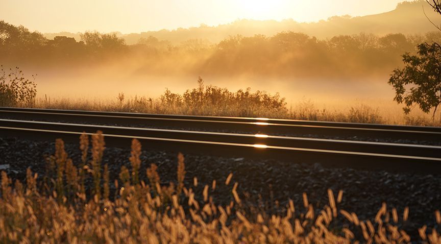 Railroad tracks in golden light, with mist over fields and trees at sunrise.