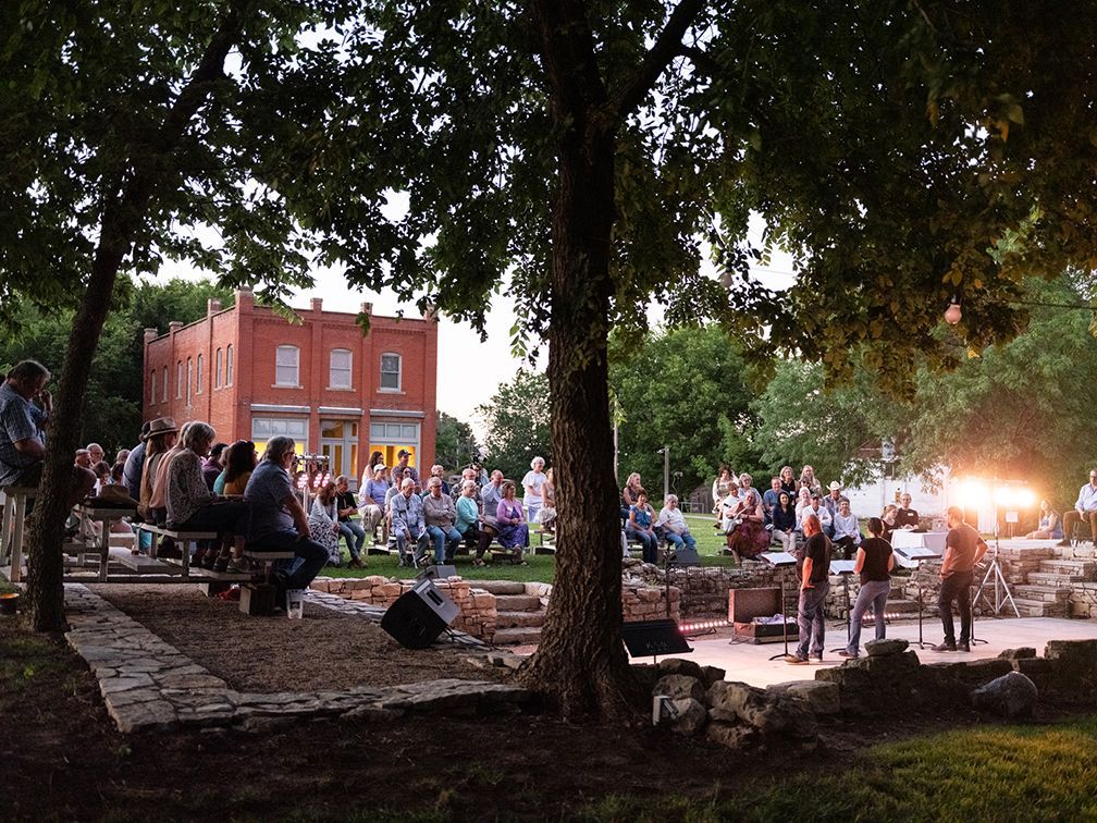 Outdoor event with a brick building in the background, people are gathered in the grassy area and near a small pond.